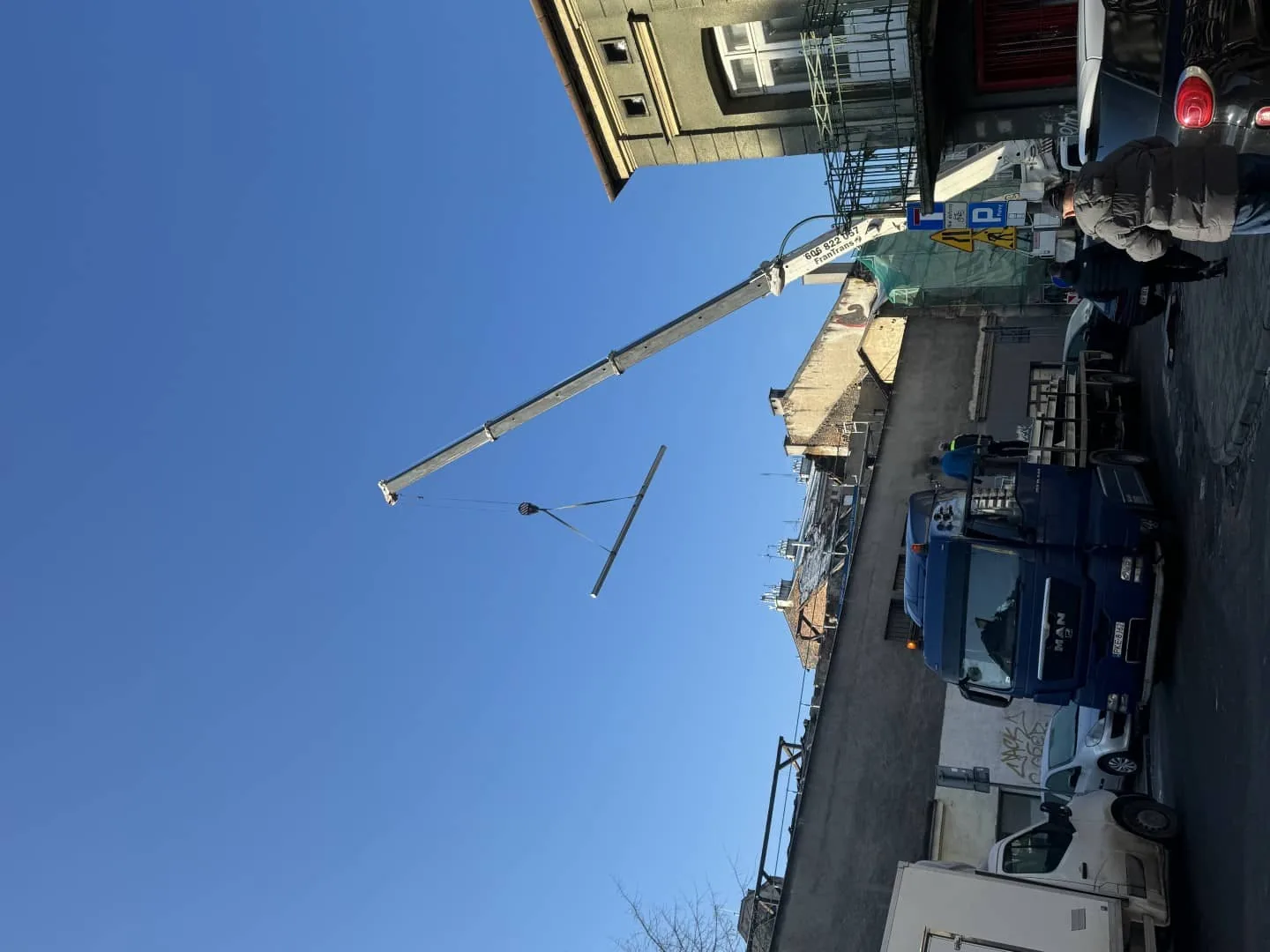 Dźwig podaje długi stalowy element nad ulicą Krakowa, obok naczepa dostawcza z konstrukcją
